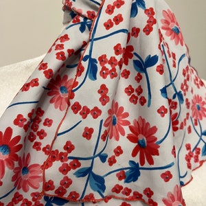 May include: A white scarf with a red and blue floral pattern. The scarf has a scalloped edge and is tied in a loose knot.