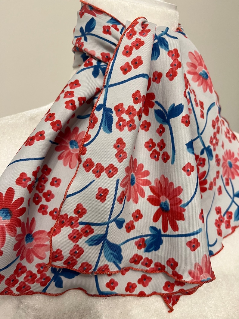 May include: A white scarf with a red and blue floral pattern. The scarf has a scalloped edge and is tied in a loose knot.