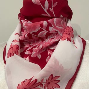 May include: A red and white floral scarf. The scarf features a vibrant red background with white and pink floral patterns. The edges are trimmed with a darker red border, creating a striking contrast. The scarf is draped, showcasing its design.