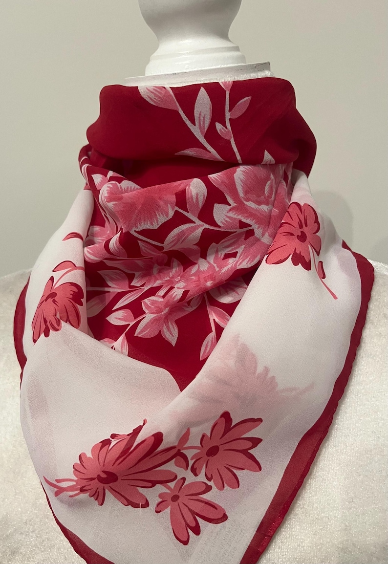 May include: A red and white floral scarf. The scarf features a vibrant red background with white and pink floral patterns. The edges are trimmed with a darker red border, creating a striking contrast. The scarf is draped, showcasing its design.