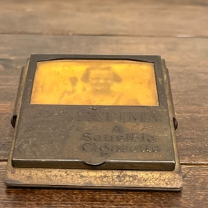 May include: Antique brass cigarette case with a hinged lid. The case features the text "FATIMA A Sensible Cigarette" and a faded image of a person. The case is rectangular and has a tarnished appearance.