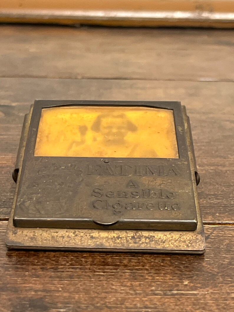 May include: Antique brass cigarette case with a hinged lid. The case features the text "FATIMA A Sensible Cigarette" and a faded image of a person. The case is rectangular and has a tarnished appearance.