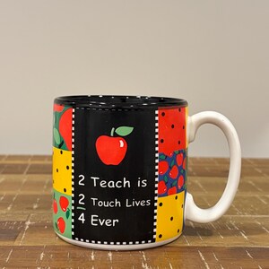 May include: A ceramic mug with a black background featuring a red apple graphic and the white text 2 Teach is 2 Touch Lives 4 Ever. The mug has a white handle and a patchwork-style border with red apple patterns and yellow polka dot sections.