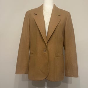May include: A tan blazer displayed on a mannequin. The blazer has a notched lapel, two front pockets, and a single button closure. The fabric appears to be a wool blend, and the color is a warm, neutral tone.