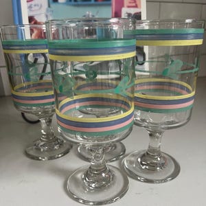 May include: A collection of clear glass goblets with colourful horizontal stripes in blue, yellow, and pink. Each goblet has a floral design and a stemmed base. The glasses are arranged on a white surface.