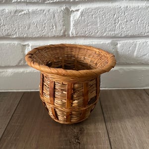 Puede incluir: Una pequeña cesta redonda tejida con un borde acampanado. La cesta está hecha de material tejido marrón claro con tiras verticales de material marrón más oscuro envueltas alrededor de los lados. La cesta está sobre una superficie de madera.