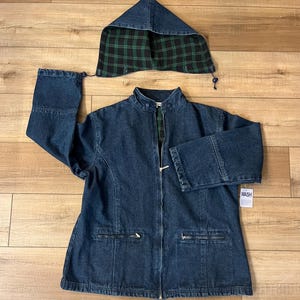 May include: A dark blue denim jacket with a matching hood. The hood's interior features a green and black plaid pattern. The jacket has a zipper closure, two front pockets, and a small tag that says "WASH".