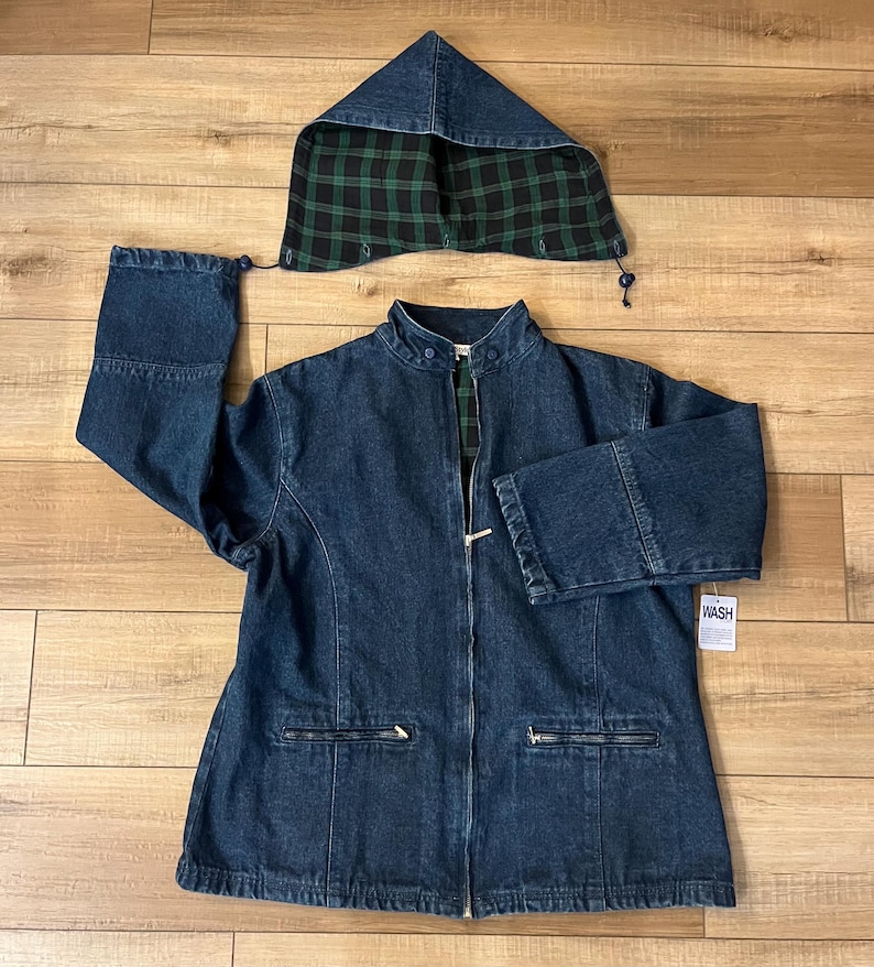 May include: A dark blue denim jacket with a matching hood. The hood's interior features a green and black plaid pattern. The jacket has a zipper closure, two front pockets, and a small tag that says "WASH".