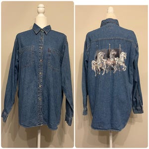 May include: A blue denim button-down shirt with long sleeves and two chest pockets. The back of the shirt features a carousel horse graphic in white, gray, and pink. The shirt is displayed on a mannequin.
