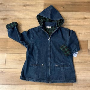 May include: A dark blue denim jacket with a hood and a full-length zipper. The hood and cuffs are lined with a green and black plaid pattern. The jacket has two front pockets and a tag that says "Wash".
