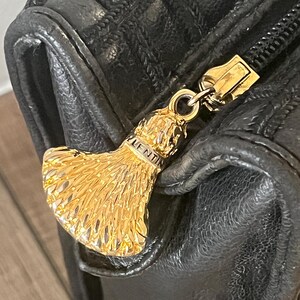 May include: A black textured leather bag featuring a gold-toned metal zipper pull shaped like a fan or tassel. The metal pull is engraved with the text VALENTIN. The bag has a quilted pattern on the upper section and a smooth leather lower section.