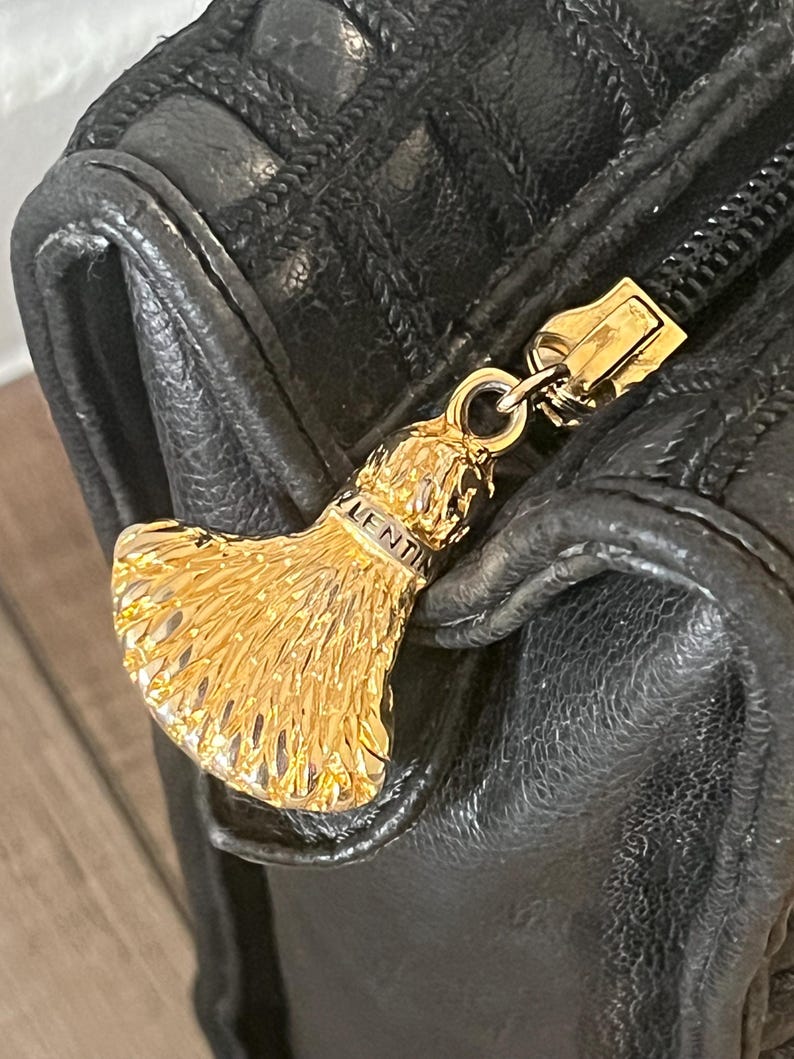 May include: A black textured leather bag featuring a gold-toned metal zipper pull shaped like a fan or tassel. The metal pull is engraved with the text VALENTIN. The bag has a quilted pattern on the upper section and a smooth leather lower section.