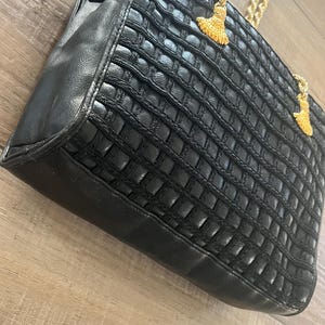 May include: A black handbag featuring a textured grid pattern on the front panel, smooth black side panels, and gold-toned metal chain straps. Two decorative gold-toned tassels are attached to the top of the bag near the chain strap connections.