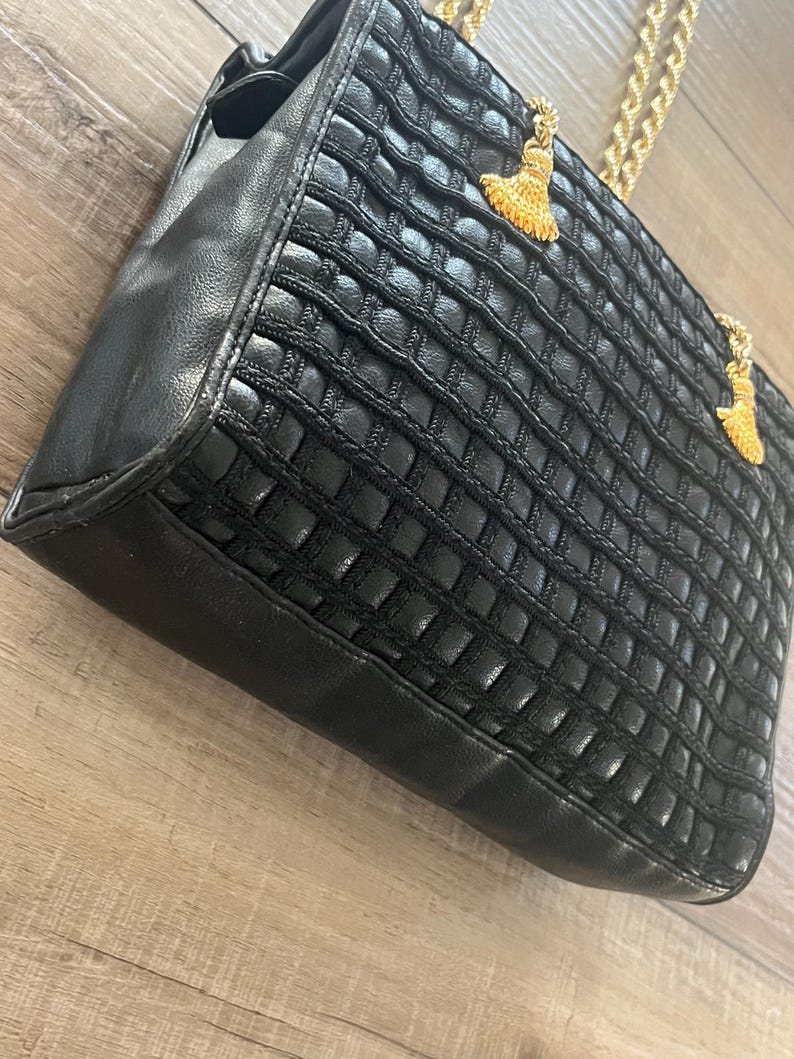 May include: A black handbag featuring a textured grid pattern on the front panel, smooth black side panels, and gold-toned metal chain straps. Two decorative gold-toned tassels are attached to the top of the bag near the chain strap connections.