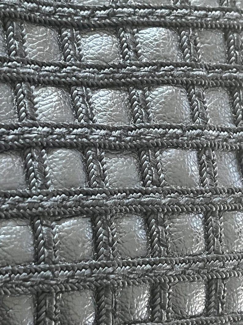 May include: A black material featuring a grid pattern created by raised, braided cord trim. The square sections between the cords have a textured, pebbled surface, creating a repeating geometric design across the entire surface of the item.