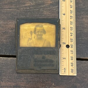 May include: Vintage metal cigarette case with a sepia-toned image of a person and the text "FATIMA A Sensible Cigarette." The case is approximately 3 inches tall, as measured by the ruler.