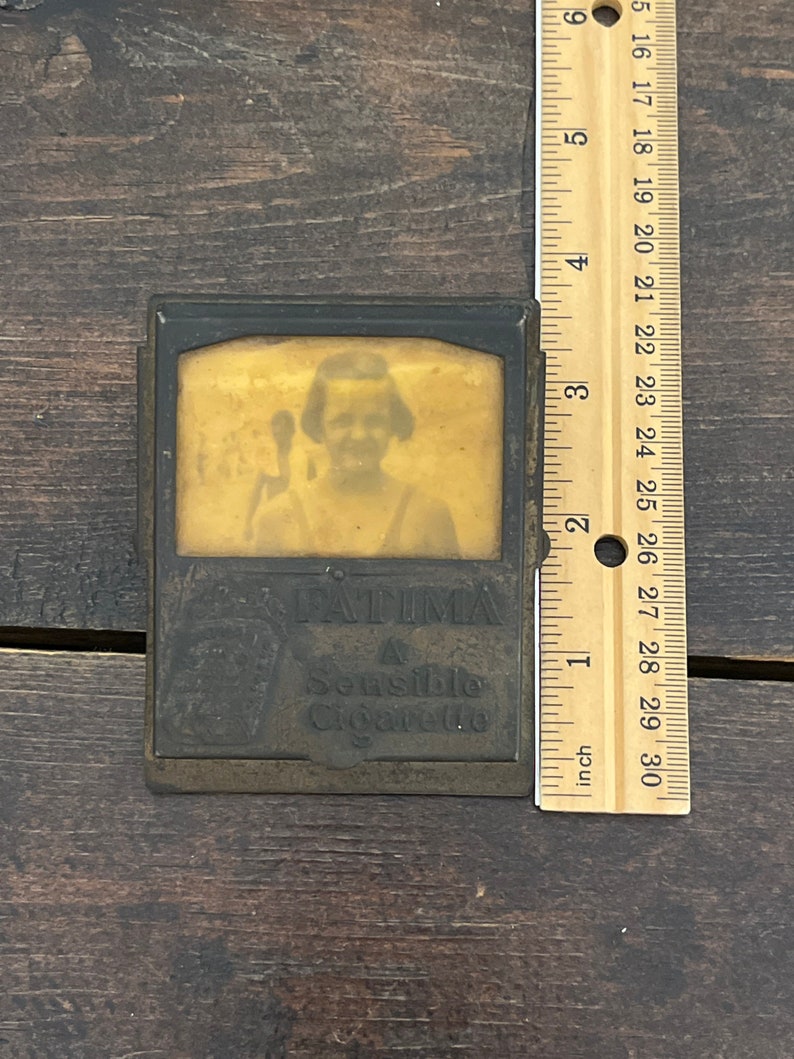 May include: Vintage metal cigarette case with a sepia-toned image of a person and the text "FATIMA A Sensible Cigarette." The case is approximately 3 inches tall, as measured by the ruler.