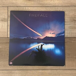 May include: A vinyl record album cover with the title "FIREFALL" in white letters. The artwork depicts a night scene with a lake, mountains, and two streaks of light across the sky. The color palette is dominated by blues and purples.