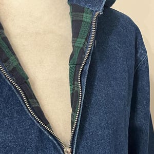 May include: Close-up of a blue denim jacket with a silver zipper. The jacket's interior lining features a green and black plaid pattern. The collar is visible, and the jacket appears to be made of a sturdy material.