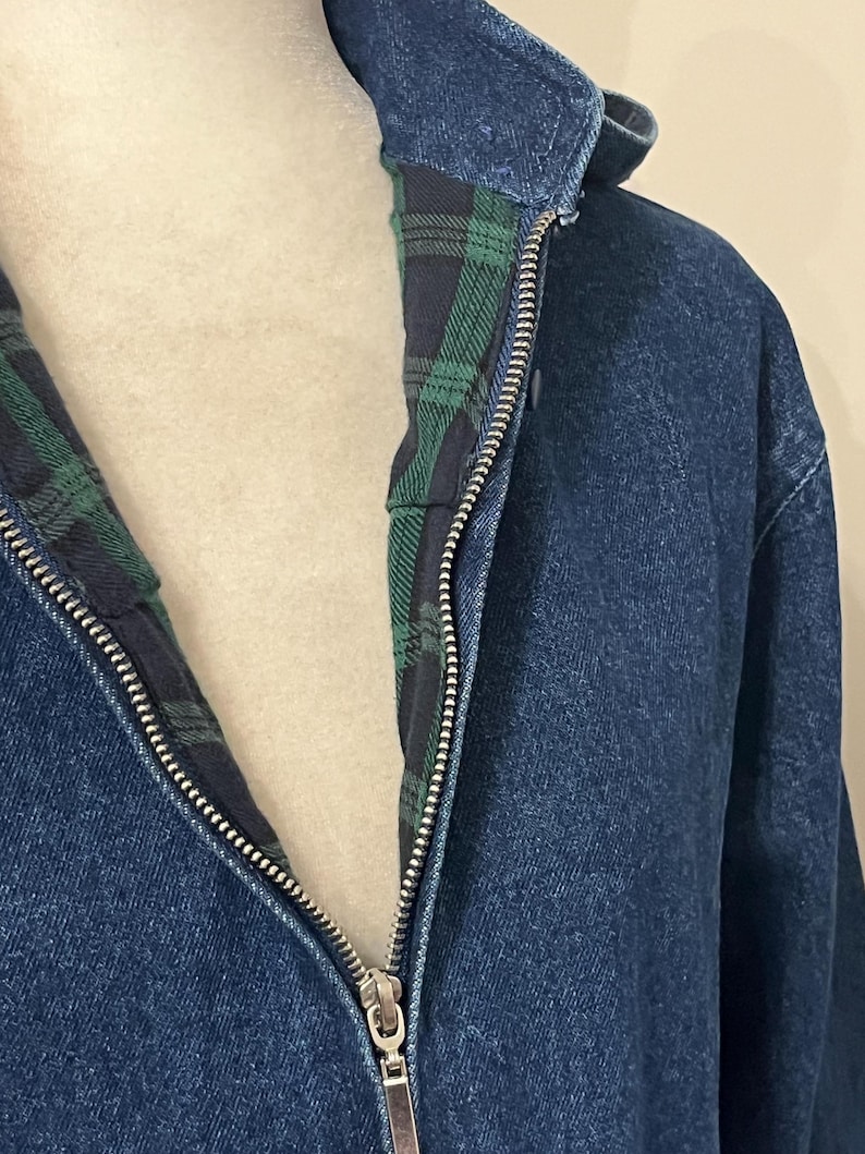 May include: Close-up of a blue denim jacket with a silver zipper. The jacket's interior lining features a green and black plaid pattern. The collar is visible, and the jacket appears to be made of a sturdy material.