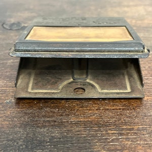 May include: An antique metal stamp holder with a rectangular window and a hinged base. The metal has a dark, aged appearance, with visible wear and tear. The window reveals a tan-colored surface, possibly for the stamp itself. The design suggests a vintage office supply.