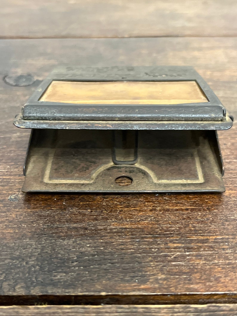 May include: An antique metal stamp holder with a rectangular window and a hinged base. The metal has a dark, aged appearance, with visible wear and tear. The window reveals a tan-colored surface, possibly for the stamp itself. The design suggests a vintage office supply.