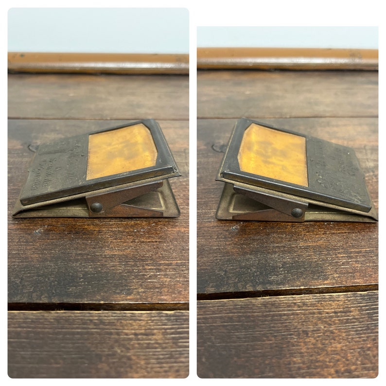 May include: Two antique metal paperweights with a hinged clip design. Each paperweight has a rectangular shape with a brass-colored insert and a dark metal frame. The clip mechanism is visible, and the paperweights are displayed on a wooden surface.