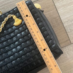 May include: A black handbag with a woven grid texture and gold-tone rope accents. A wooden ruler is placed across the bag, showing a length of approximately 9.5 inches. The bag features a gold-tone tassel detail and a structured rectangular shape.