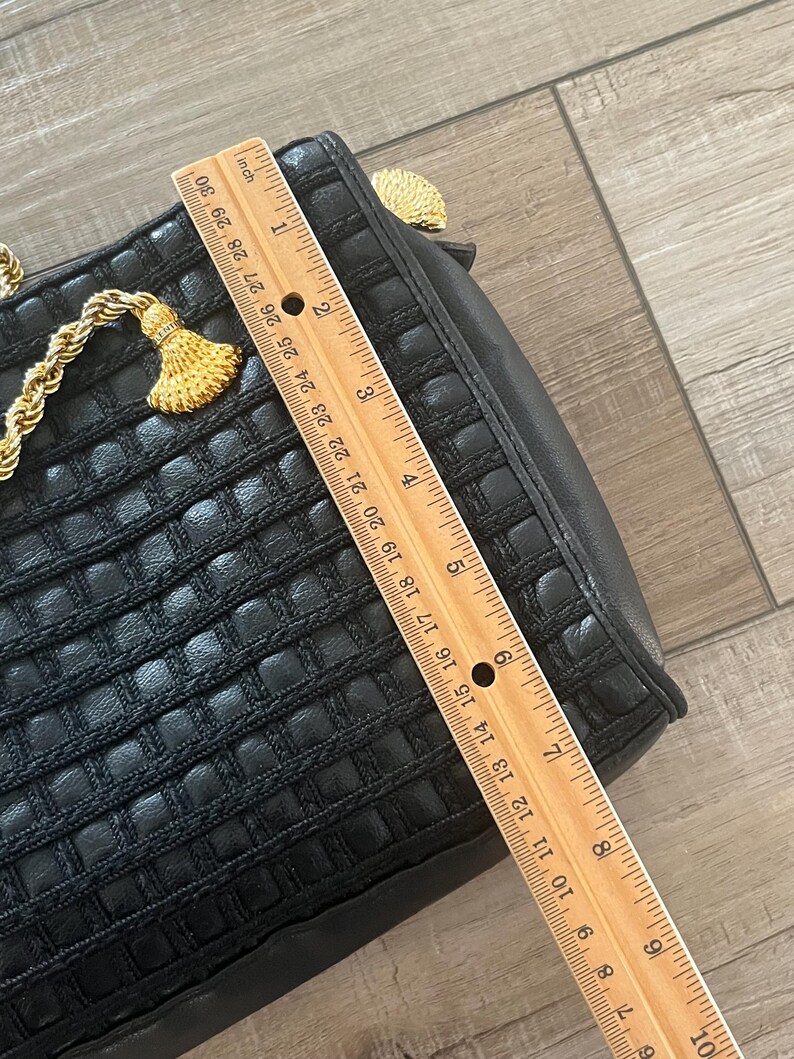 May include: A black handbag with a woven grid texture and gold-tone rope accents. A wooden ruler is placed across the bag, showing a length of approximately 9.5 inches. The bag features a gold-tone tassel detail and a structured rectangular shape.