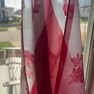 May include: A sheer white and red floral curtain hangs in front of a window. The curtain features a repeating pattern of pink flowers and geometric shapes. The sunlight shines through the fabric, creating shadows.