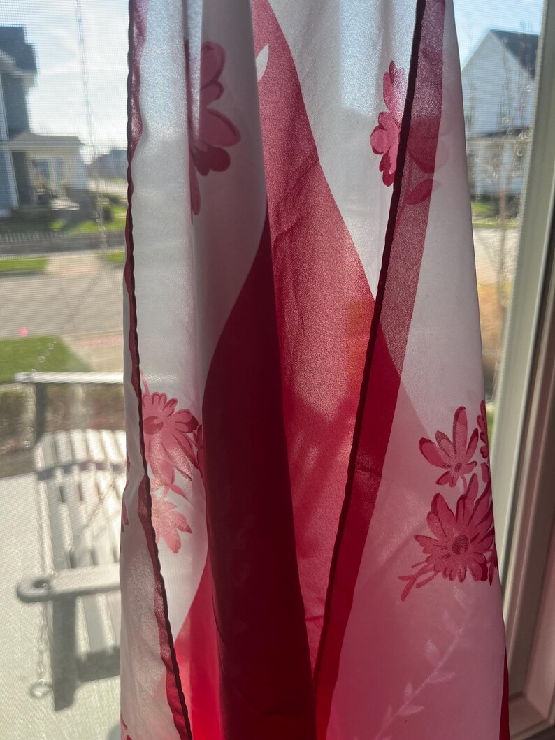 May include: A sheer white and red floral curtain hangs in front of a window. The curtain features a repeating pattern of pink flowers and geometric shapes. The sunlight shines through the fabric, creating shadows.