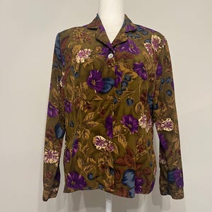 May include: A long-sleeved button-down shirt with a floral pattern. The shirt features a dark olive green background with purple, white, and blue flowers and leaves. The shirt is displayed on a mannequin.