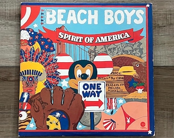 Peut inclure: Une pochette d'album vintage pour "Spirit of America" des Beach Boys. La couverture présente un collage avec Mickey Mouse, un gant de baseball, un aigle et le texte "ONE WAY". Le design incorpore des éléments rouges, blancs et bleus.