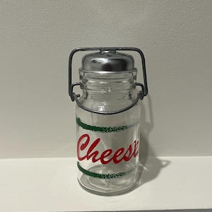 Puede incluir: Un agitador de queso de vidrio transparente con tapa y asa de metal. La palabra "Cheese" está escrita en rojo con acentos verdes. El agitador está diseñado para contener y dispensar queso.