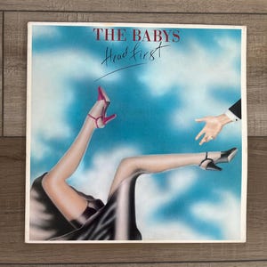 May include: A vintage vinyl record album cover featuring a woman in a black dress and red heels falling headfirst towards a hand reaching out from the clouds. The album title is "THE BABYS Head First".