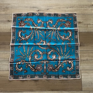 Puede incluir: Un pañuelo de seda azul cuadrado con un borde blanco y un patrón de paisley en tonos de rojo, marrón y dorado.