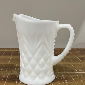 May include: A white, vintage-style pitcher with a diamond pattern and a textured handle. The pitcher has a curved spout and a round base. The pitcher is made of opaque white glass and sits on a wooden surface.