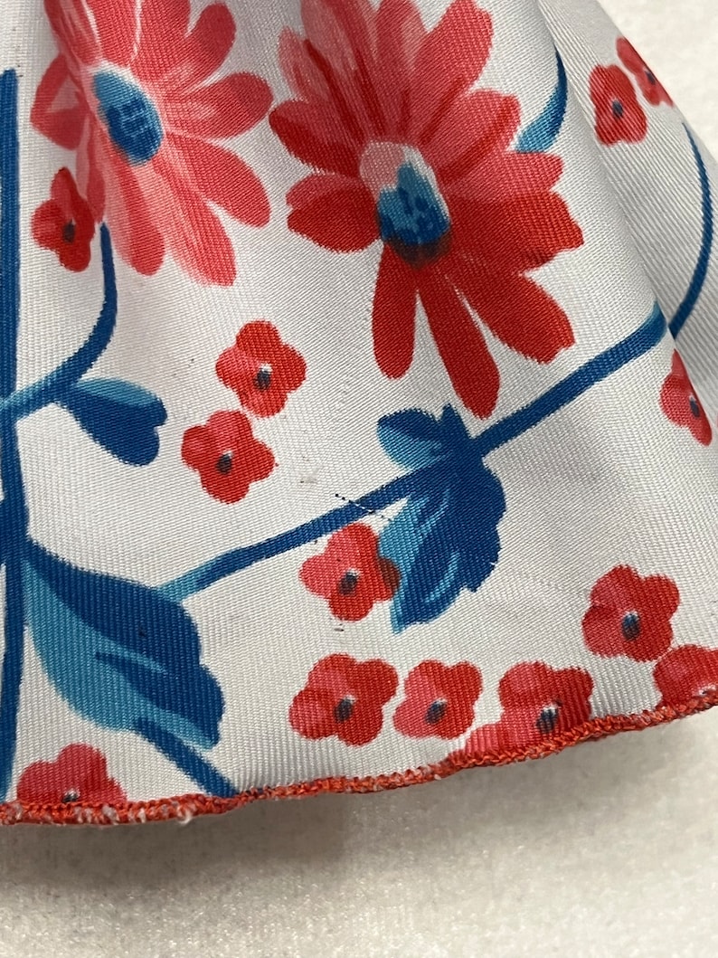 May include: A white fabric with a red and blue floral pattern. The fabric has a red trim around the edge.