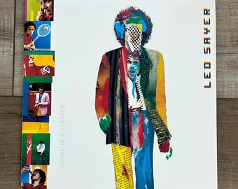 Può includere: Copertina di un album in vinile vintage per "Living in a Fantasy" di Leo Sayer. La copertina presenta un ritratto in stile collage con colori vivaci, tra cui blu, rosso, giallo e nero. Il nome dell'artista è visualizzato verticalmente in lettere colorate.