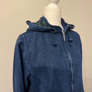 May include: A blue denim jacket with a hood. The jacket has a zipper closure and blue ball-shaped drawstrings. The hood lining features a green and black plaid pattern. The jacket is displayed on a white mannequin.
