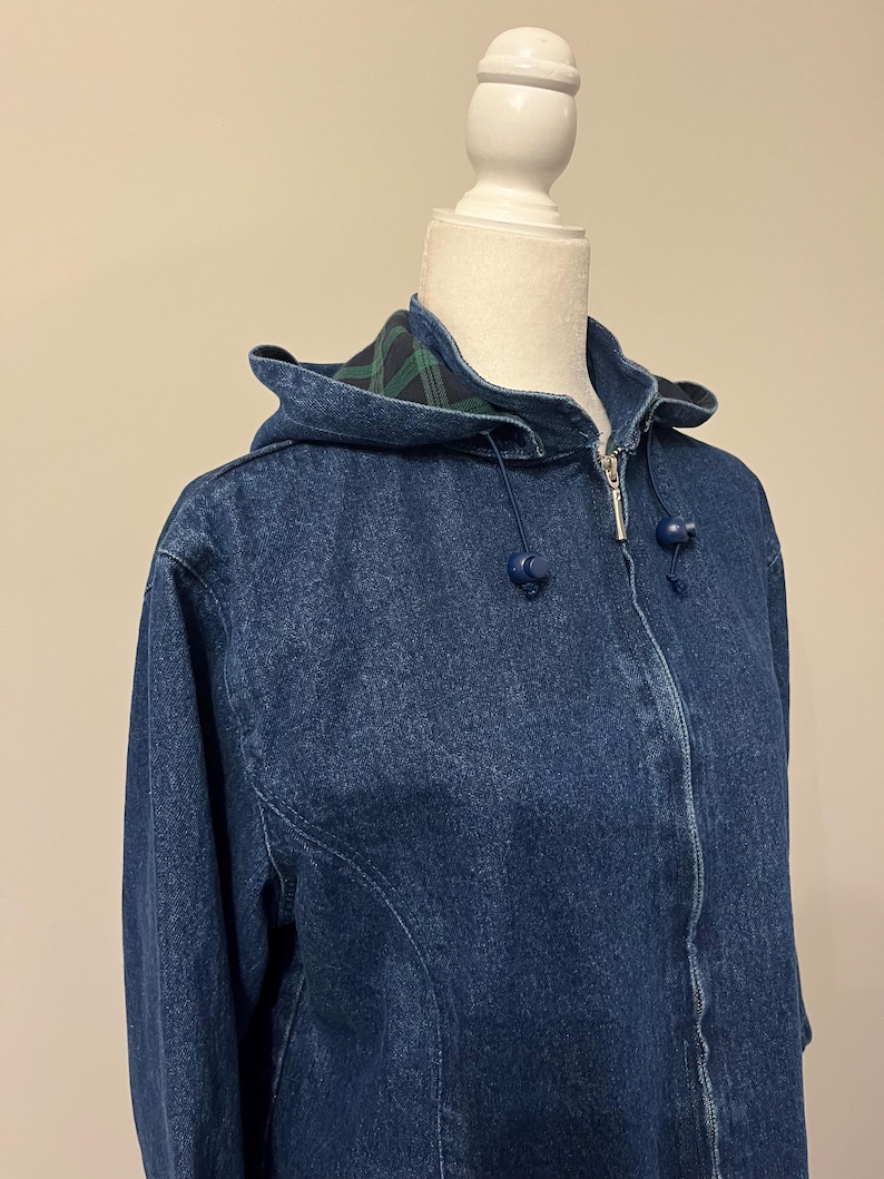 May include: A blue denim jacket with a hood. The jacket has a zipper closure and blue ball-shaped drawstrings. The hood lining features a green and black plaid pattern. The jacket is displayed on a white mannequin.