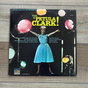 Peut inclure: Pochette d'album vinyle vintage pour "This Is Petula Clark !" avec une photo de Petula Clark en robe bleue. La pochette de l'album a un fond noir avec des orbes colorés et le logo Sunset Records.