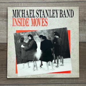 May include: Album cover for the Michael Stanley Band's "Inside Moves." The album art features a black and white photo of several people in coats, set against a red and white background. The band's name and album title are in black and red text.