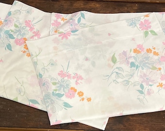 May include: Three white cotton pillowcases with a floral pattern featuring pink, blue, and orange flowers. The pillowcases are folded and stacked on a wooden surface.