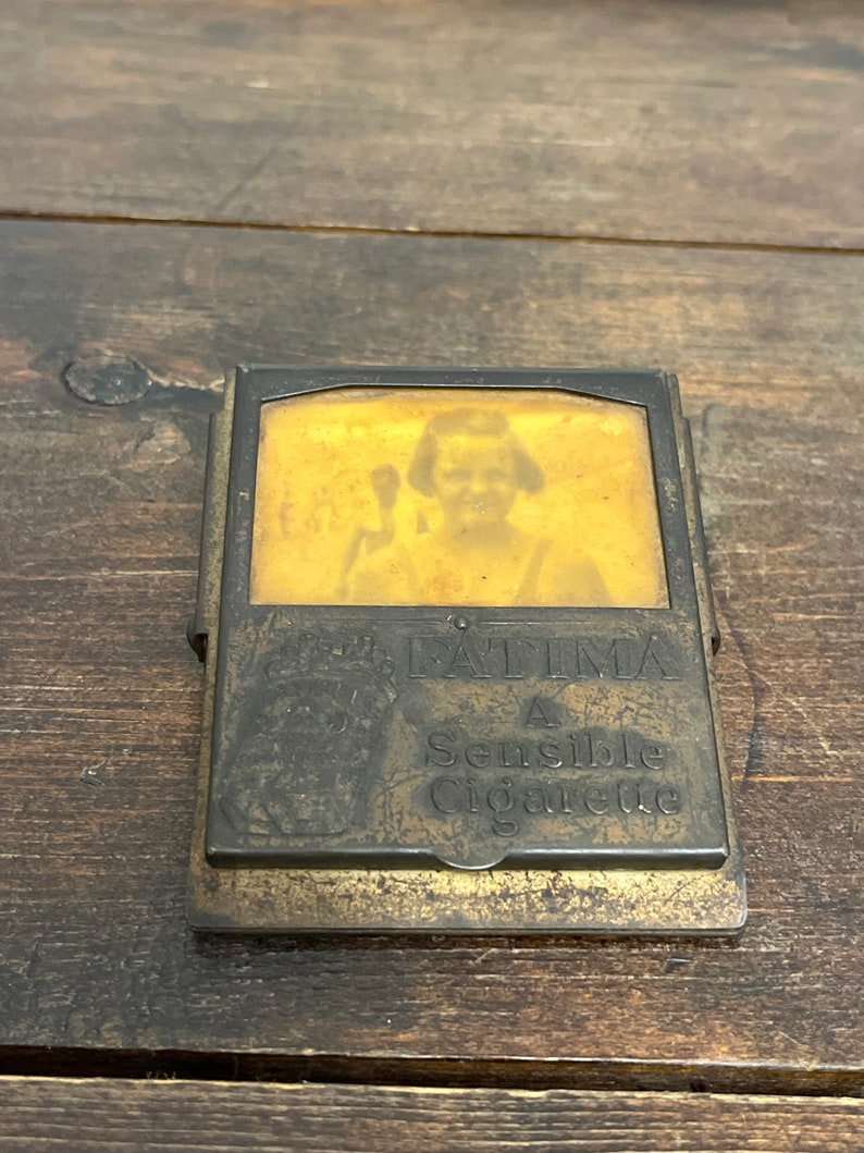 May include: Vintage brass cigarette case with a portrait and the text "FATIMA A Sensible Cigarette." The case has a rectangular shape with a hinged lid and a clasp. The brass has a worn, aged appearance.