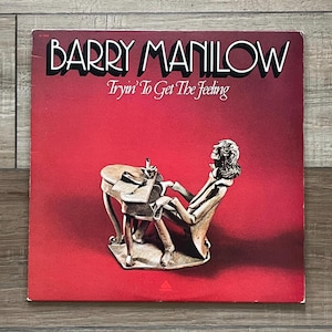 May include: A vintage vinyl record album cover for Barry Manilow's album, "Tryin' To Get The Feeling." The cover features a red background with the artist's name in white and the album title in a cursive font. A sculpture of a person at a table is in the foreground.
