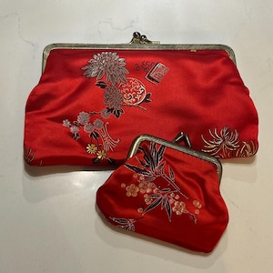 May include: Two red satin clutch bags with floral embroidery. The larger bag has a rectangular shape with a metal clasp, and the smaller bag is a coin purse. Both bags feature gold and silver floral designs.