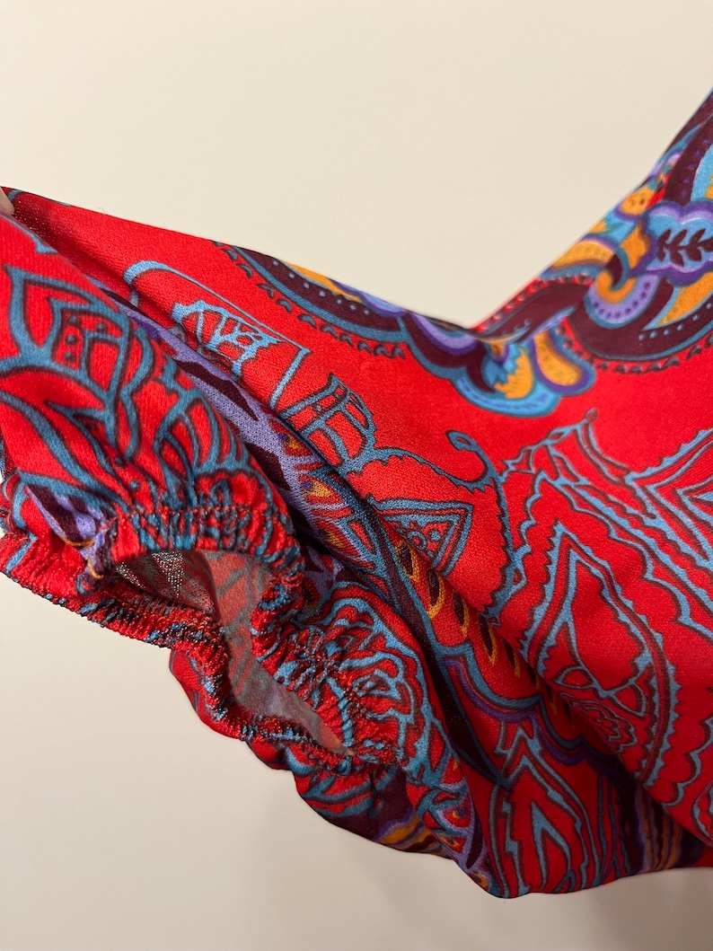 May include: Red fabric with a blue, purple, and yellow paisley pattern. The fabric has a gathered edge.
