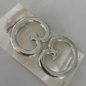 May include: Two silver-toned metallic circular fasteners with an open center and a stylized inward-pointing hook detail. The items are attached to a white rectangular display card featuring the brand name Streamline printed in black ink.