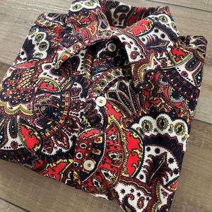 May include: A folded button-down shirt with a vibrant paisley pattern in red, black, yellow, and white. The shirt features a classic collar and three visible white buttons. The fabric appears to be a woven material.